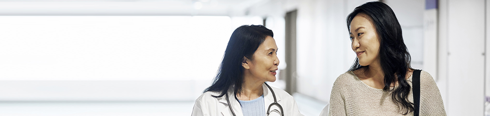 Smiling mature doctor talking to woman in hospital. Female patient visiting healthcare worker for routine checkup. They are standing in corridor.